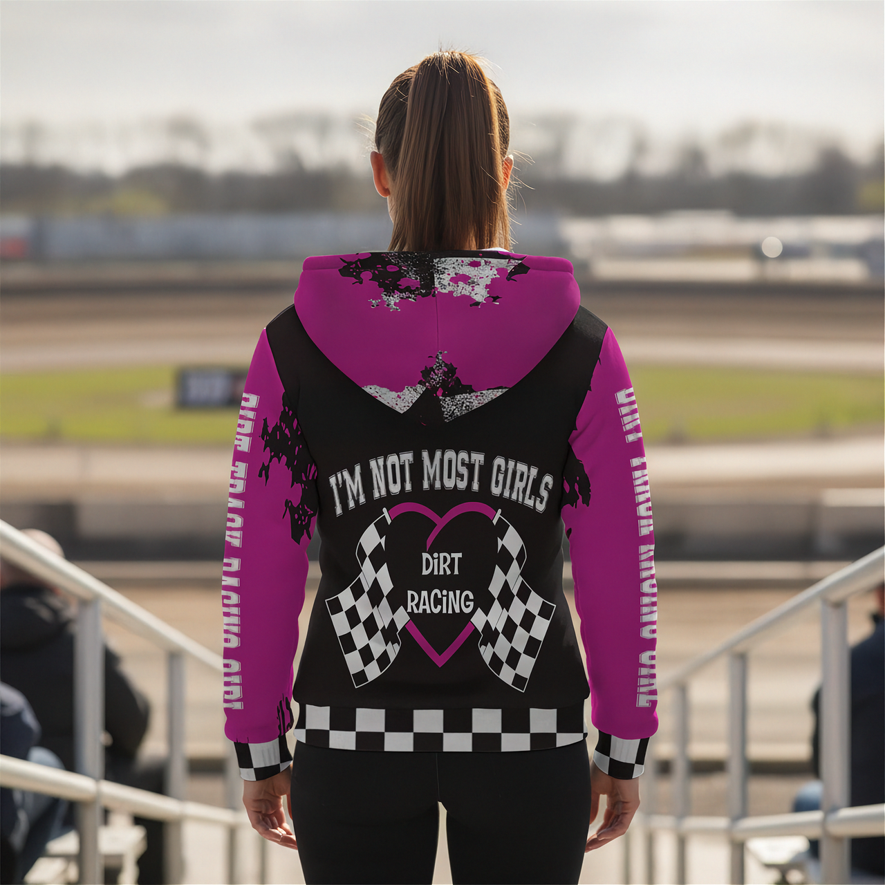 Woman wearing a pink and black hoodie with 'I'm Not Most Girls Dirt Racing' text at a race track.
