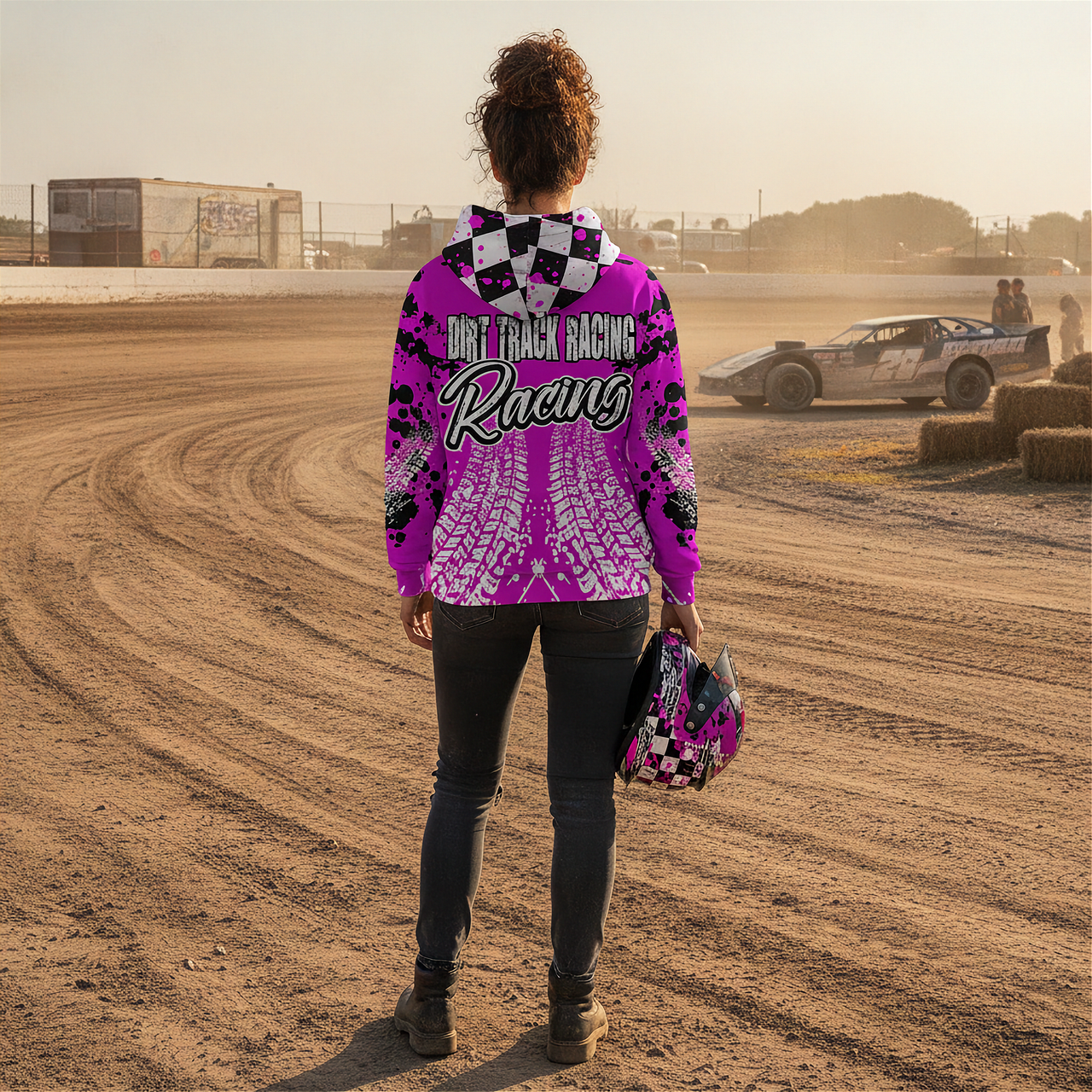 Woman wearing a pink 'Dirt Track Racing' hoodie on a dirt track with a car in the background.