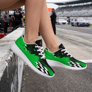 A pair of low-top racing-inspired sneakers worn on a person's feet, photographed at a racetrack. The shoes are bright green and black with white laces, white soles, and a black-and-white checkered pattern near the front.