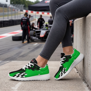 A person wearing bright green and black checkered sneakers with white soles, sitting on a concrete barrier at a racetrack while wearing gray leggings. An F1 race car and track environment are blurred in the background.