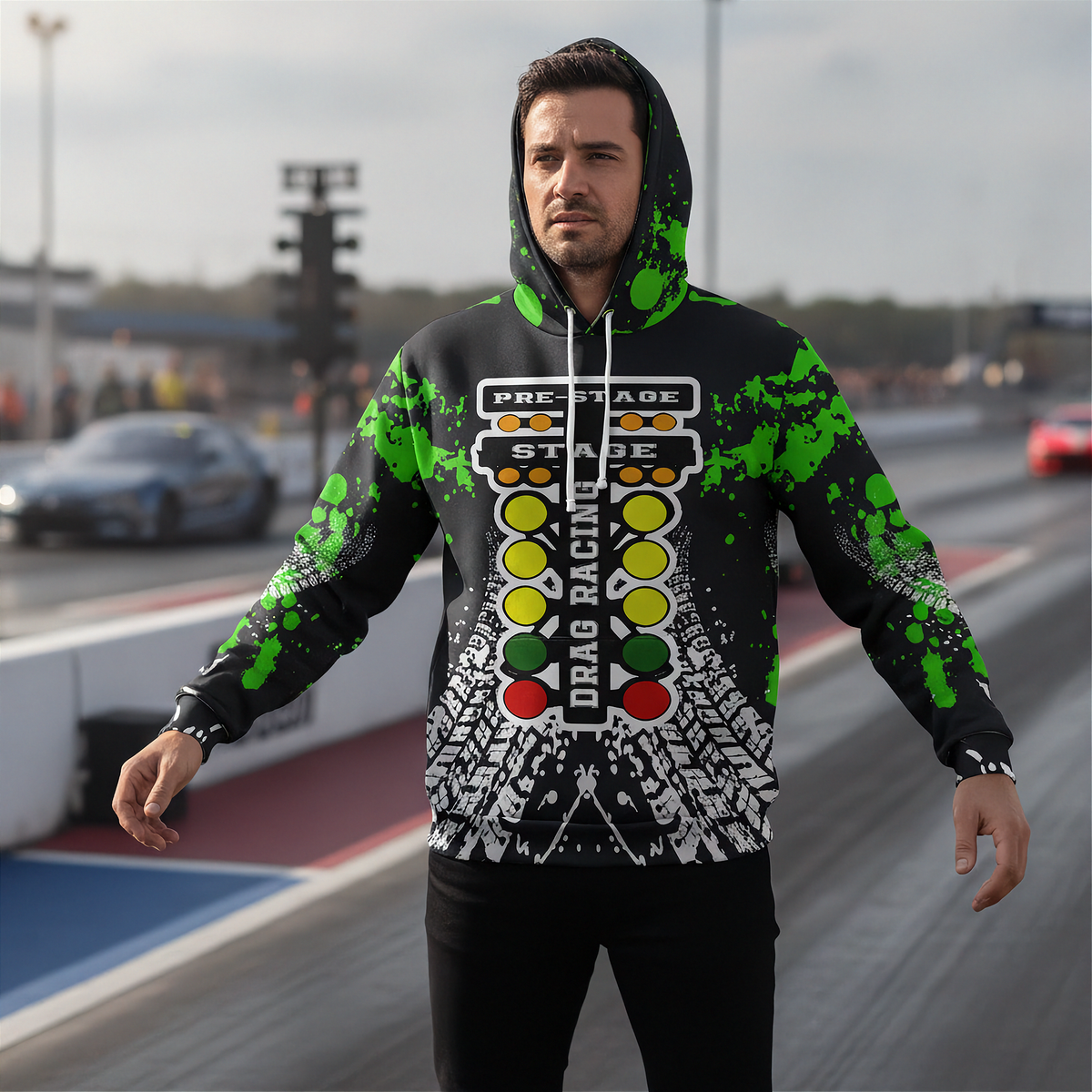 A man wearing a black hoodie with a green, yellow, red, and white drag racing theme stands on a racetrack. The hoodie features a drag racing light column graphic and green paint splatters on the sleeves and hood.