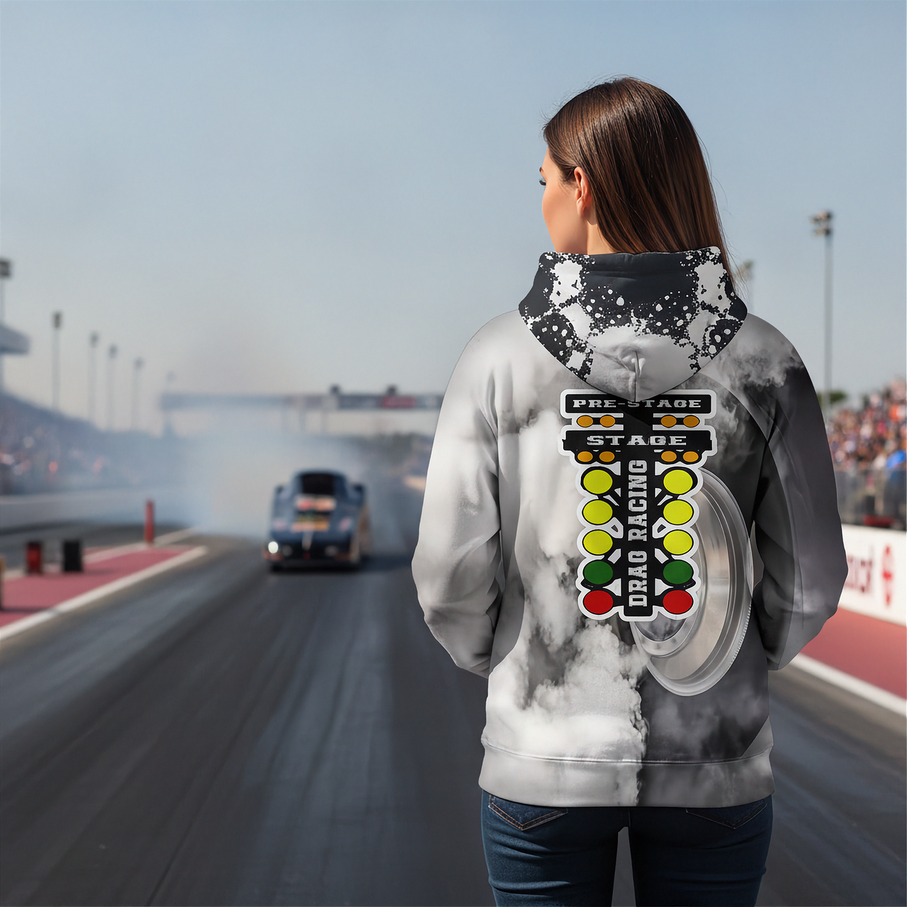 A woman stands on a drag racing track wearing a white and gray smoky hoodie with a large drag racing graphic on the back, including a start line and traffic light design. A race car is visible in the distance on the track.