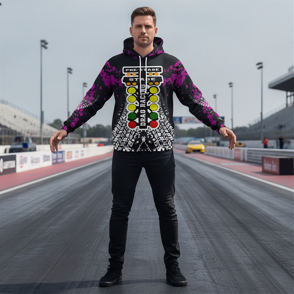 A man stands on a racetrack wearing a black hoodie with a colorful racing-inspired graphic print featuring a vertical slot machine design and purple sleeve patterns, paired with black pants.