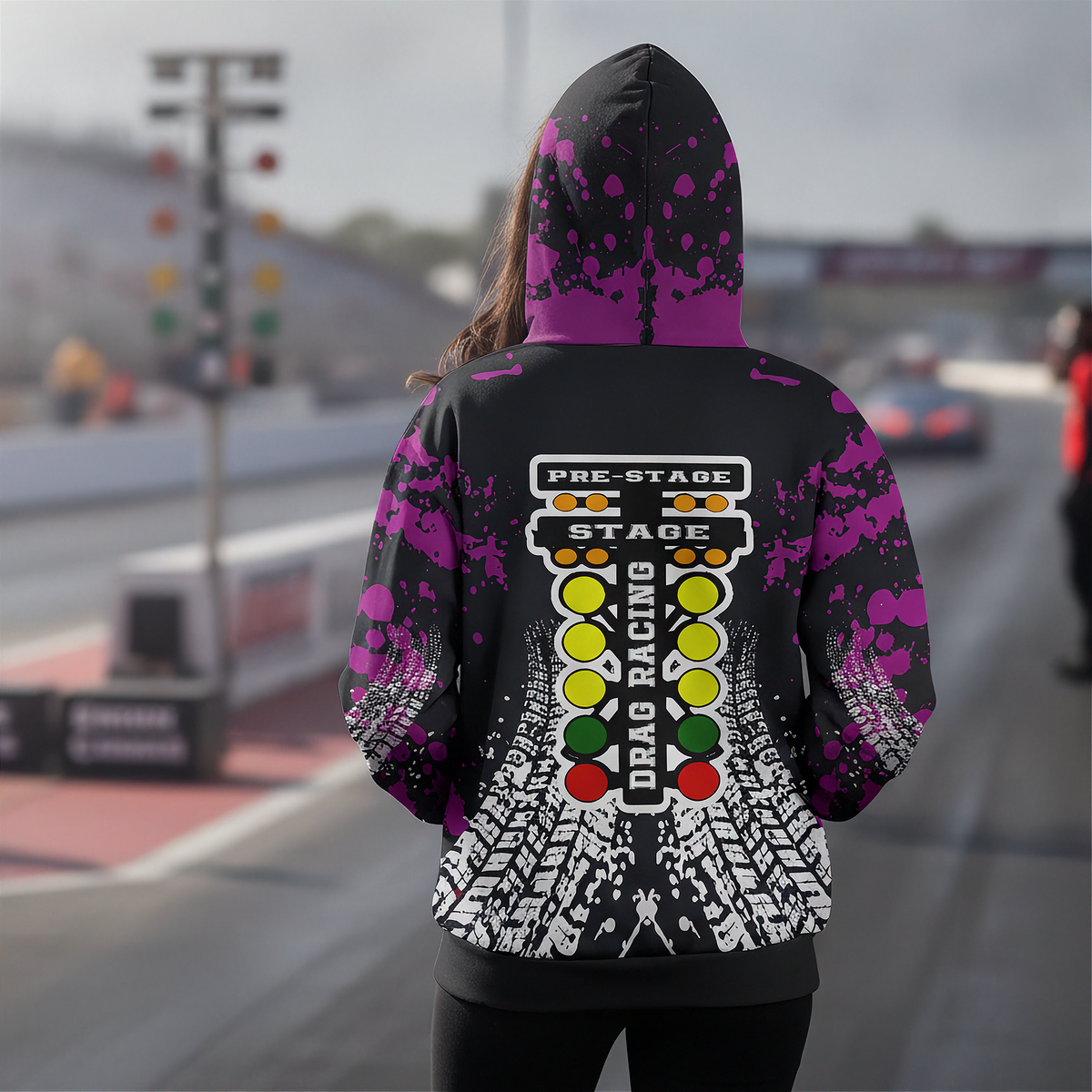A person standing on a racetrack wearing a black hoodie with a large drag racing graphic on the back, featuring a start bar design and tire tracks, with purple paint splatter details on the sleeves and hood.