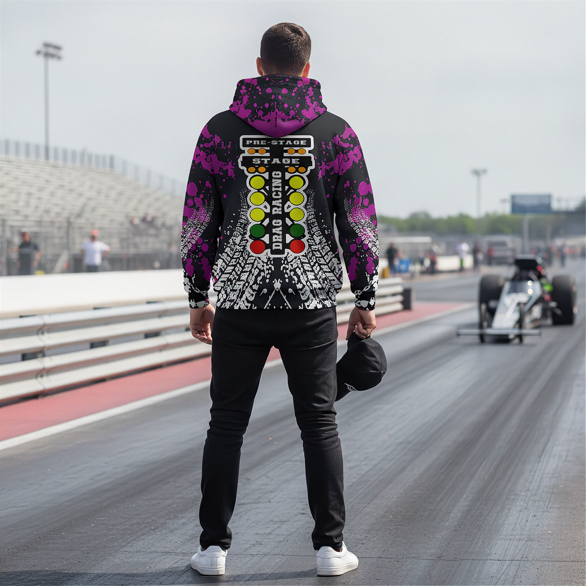 A man stands on a drag racing track wearing a black hoodie with a purple and white graphic design featuring a race track flag pattern on the back. In the background, a drag racer is visible on the track.