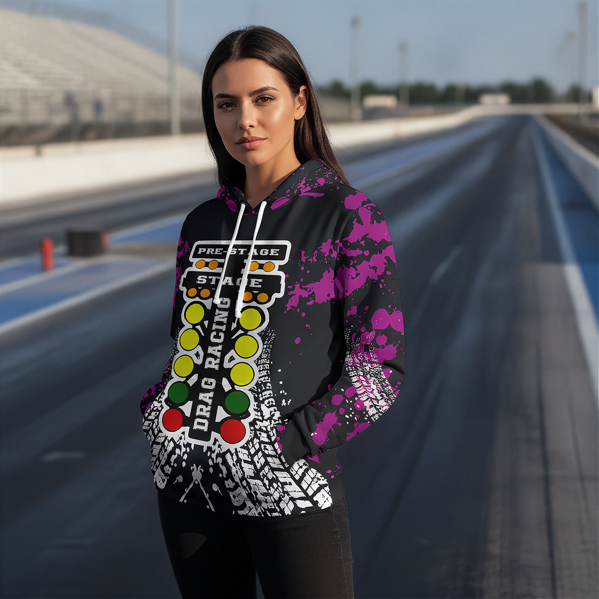 A woman stands on a racetrack wearing a black hoodie with a drag racing graphic print featuring purple paint splatters, a checkered flag design, and colorful start-line buttons.