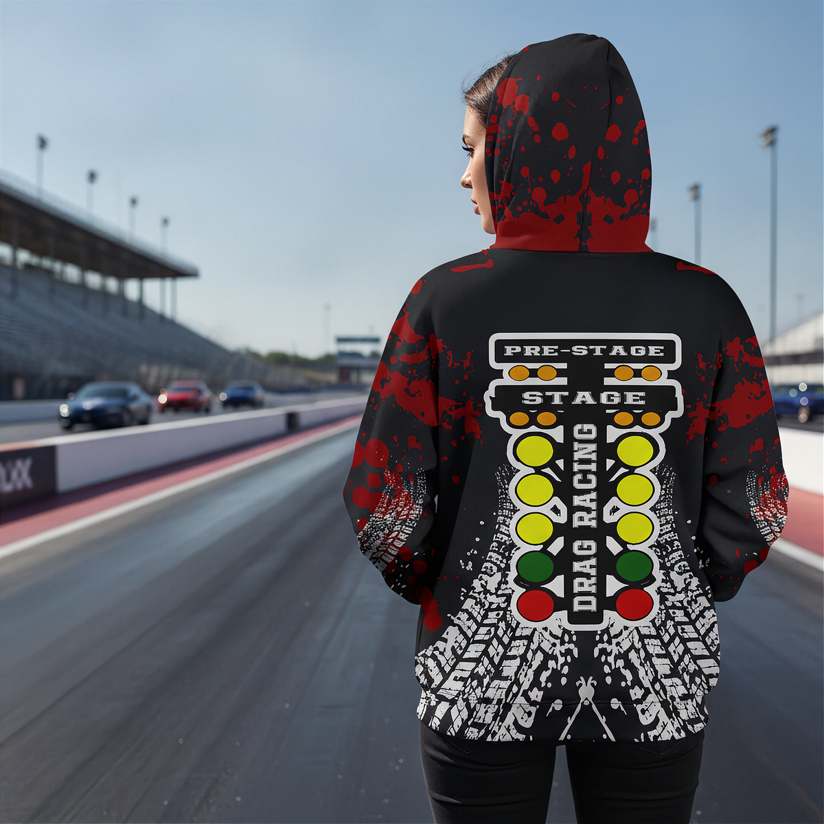 A person standing on a racetrack wearing a black hooded sweatshirt with a red splatter pattern and a large drag racing stage graphic on the back, shown from behind.