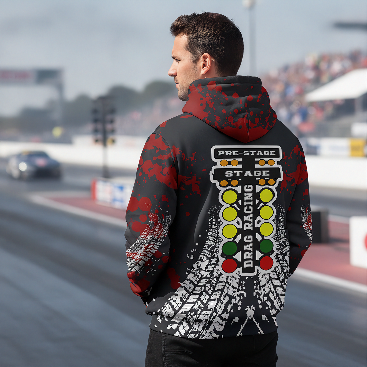 A man stands on a racetrack wearing a black hoodie with a red hood and a large drag racing graphic on the back, featuring a light bar, racing lights, and tire tracks.