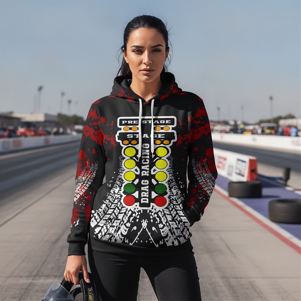 A woman stands on a racetrack wearing a black hoodie with a red, white, and yellow drag racing graphic print, paired with black leggings.
