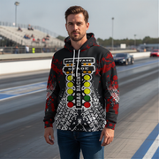 A man wearing a black, red, and white drag racing themed hoodie stands on a racetrack with grandstands and cars in the background.