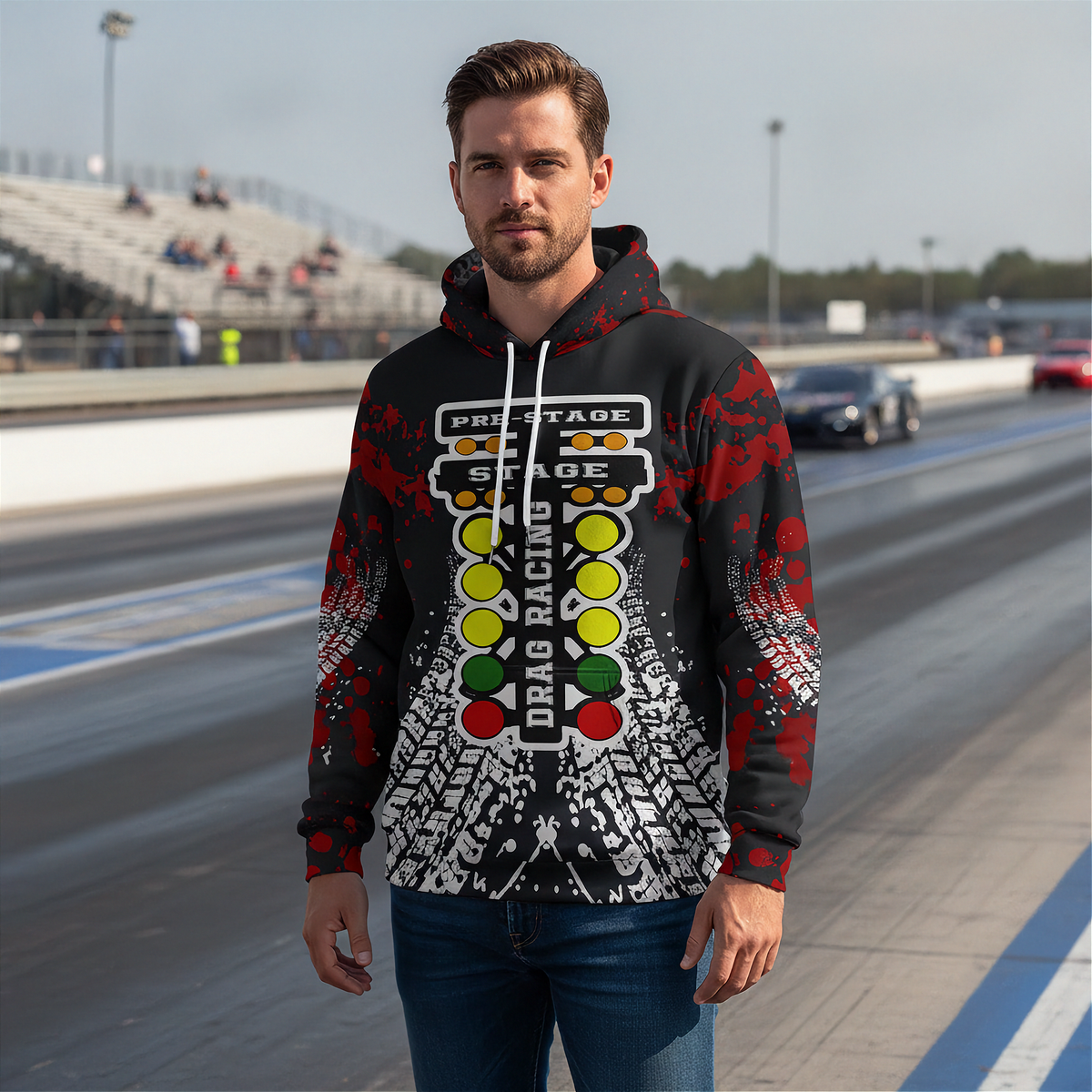 A man wearing a black, red, and white drag racing themed hoodie stands on a racetrack with grandstands and cars in the background.
