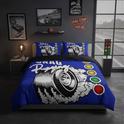 A racing-themed bedding set featuring a blue duvet cover and matching pillowcases with a large black-and-white tire graphic, smoke, and colorful drag strip light buttons, styled in a dark gray bedroom with racing decor.
