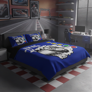 A racing-themed bedding set with a blue comforter and matching pillowcases featuring a large black-and-white driver graphic and colorful oval accents, styled in a modern gray gaming room with a desk, chair, shelves, and checkered floor.