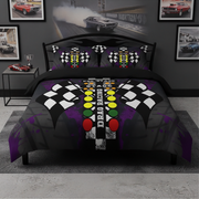 A drag racing themed bedding set featuring a duvet cover and two pillowcases with a black and purple design, checkered flags, a traffic light motif, and the words 'DRAG RACING'. The set is displayed on a bed in a dark gray bedroom decorated with framed drag race posters.