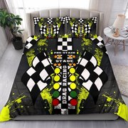 A bed dressed in a drag racing themed bedding set, featuring a duvet cover and two matching pillowcases with a black and white checkered flag design, neon yellow paint splatters, and a traffic light graphic. The bed is styled in a modern bedroom setting with a headboard, side table, lamp, and plant.