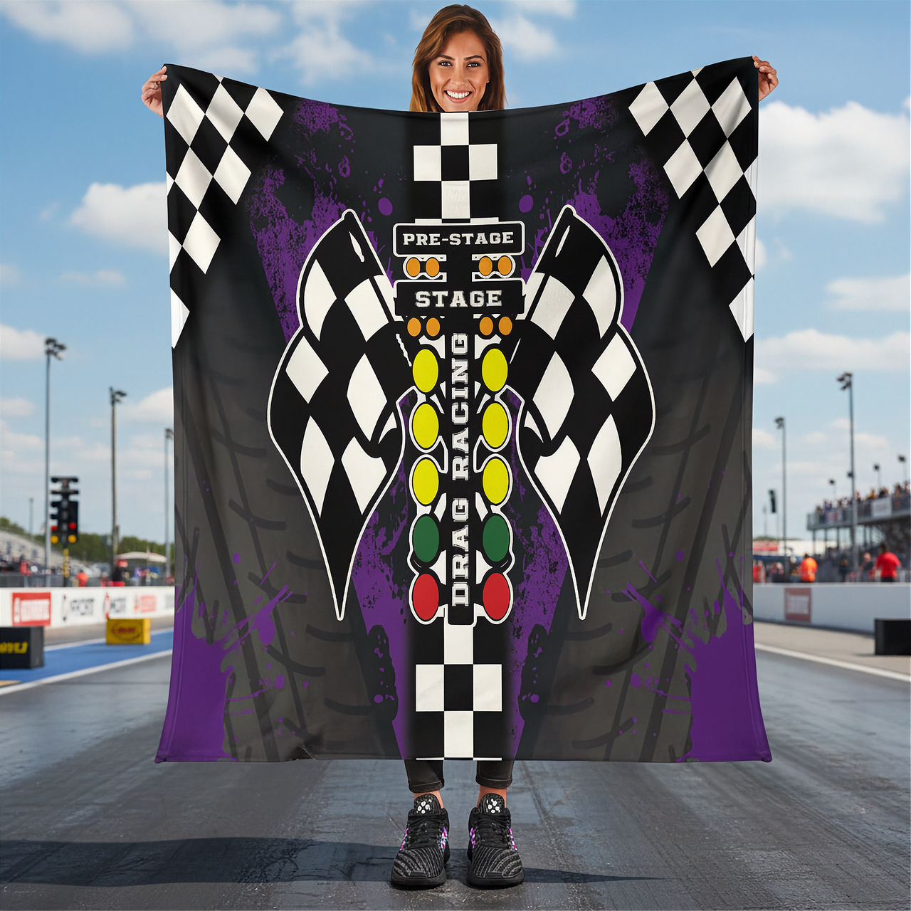 Person holding a drag racing themed blanket with checkered flags and race track elements at a race track.