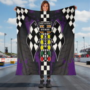 Person holding a drag racing themed blanket with checkered flags and race track elements at a race track.
