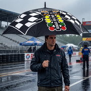 A man standing on a wet racetrack under a large black racing umbrella with a checkered flag and traffic light graphic, wearing a dark rain jacket. Race track, grandstands, and other umbrellas are visible in the background.