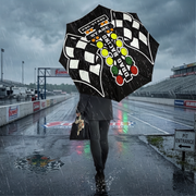 A person walking on a wet racetrack in the rain, holding a large black umbrella printed with a checkered flag pattern, 'Tibet Racing' text, and colorful oval logos. Race track barriers, grandstands, and signage are visible in the background.