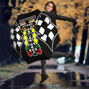 A woman jumps in a puddle outdoors while holding up a large black umbrella printed with a checkered flag design, racing lights, and the words 'PRE-STAGE DRAG RACING STAGE'.
