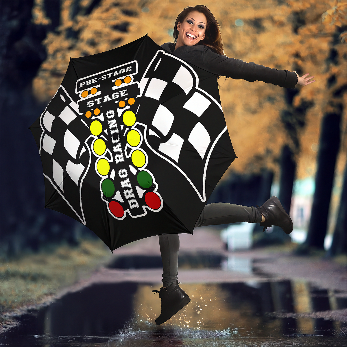 A woman jumps in a puddle outdoors while holding up a large black umbrella printed with a checkered flag design, racing lights, and the words 'PRE-STAGE DRAG RACING STAGE'.