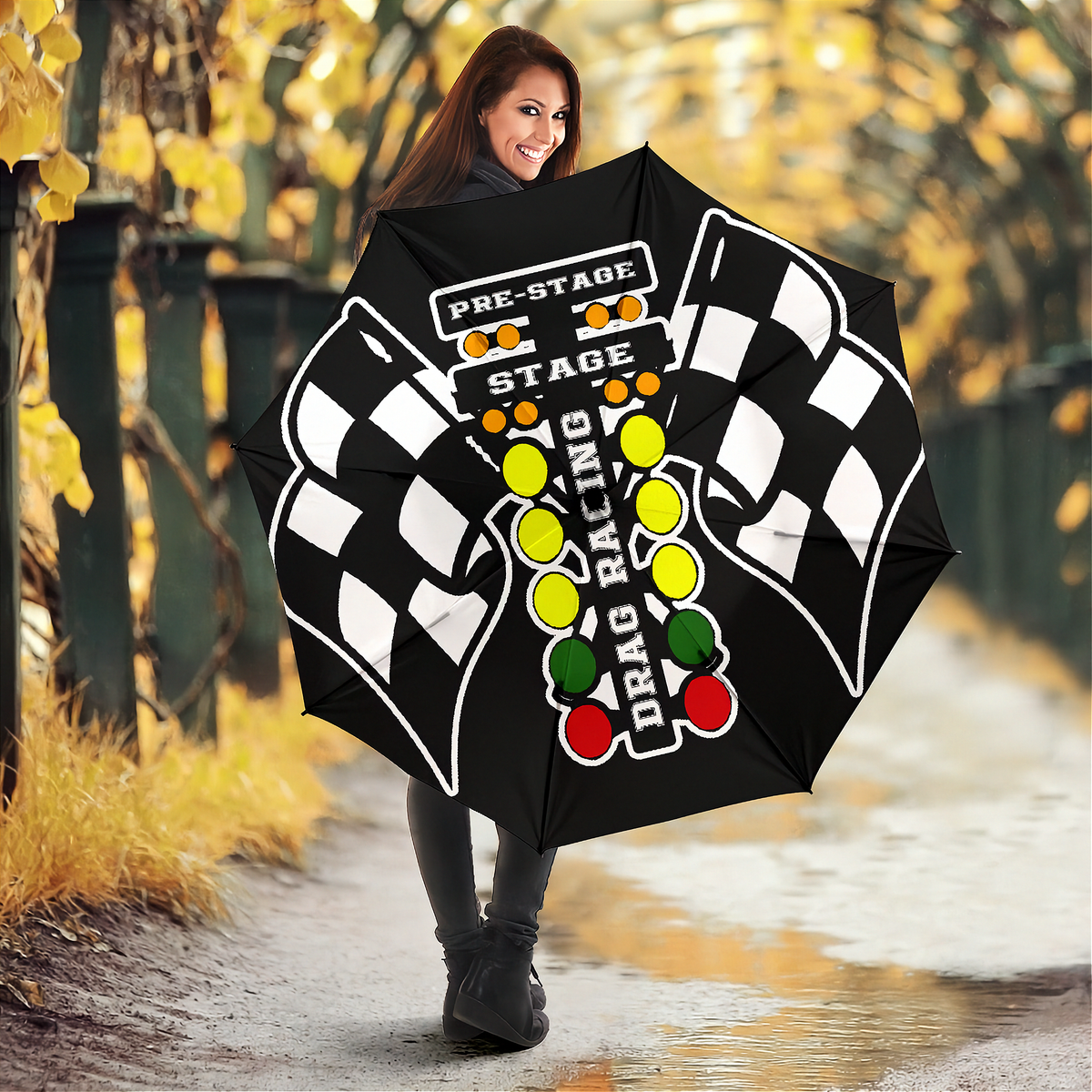A woman holding a large black racing-themed umbrella on a wet path with autumn trees in the background. The umbrella features a checkered flag pattern, the words 'PRE-STAGE' and 'STAGE', and a vertical 'DRAG RACING' graphic with green, yellow, and red lights.