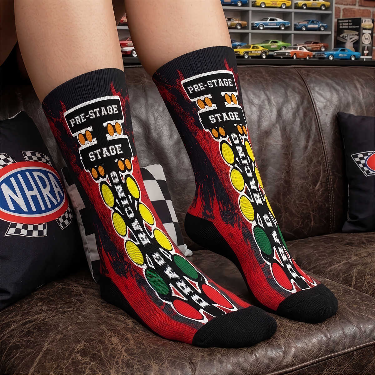 A pair of crew socks worn on a person's legs, featuring a red, black, yellow, and green racing-themed graphic with 'PRE-STAGE' and 'STAGE' text, set against a brown leather couch background.