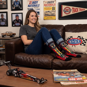 A woman sits on a brown leather couch wearing colorful NHRA-themed socks, with an NHRA-branded pillow on the couch and an NHRA poster on the wall behind her. A model drag racing car is on the table in the foreground.