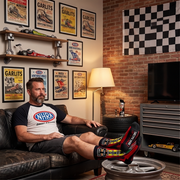 A man relaxes on a dark leather couch in a garage-themed room, wearing a black and white NHRA crew-neck t-shirt and matching DRAG RACING crew socks with colorful graphics. The background features a brick wall, a checkered flag, framed vintage race car posters, and a gray metal tool chest.