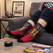 A person reclining on a gray couch with their legs crossed, wearing red and black crew socks with a racing-themed graphic design. The background shows a framed NHRA poster on a side table, a small model hot rod, and a magazine on a coffee table in the foreground.