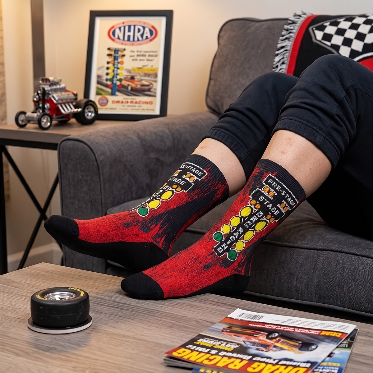A person reclining on a gray couch with their legs crossed, wearing red and black crew socks with a racing-themed graphic design. The background shows a framed NHRA poster on a side table, a small model hot rod, and a magazine on a coffee table in the foreground.