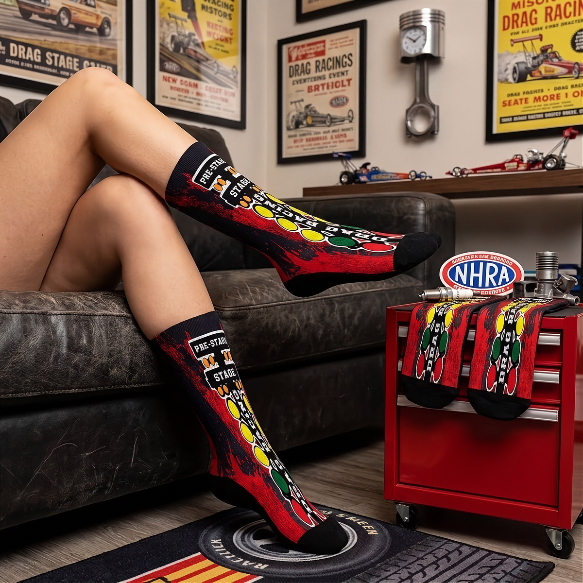 A person reclining on a dark leather couch wearing red, black, yellow, and green drag racing themed crew socks. The room background features vintage drag racing posters, a wall-mounted piston, and a red tool cart with matching socks.