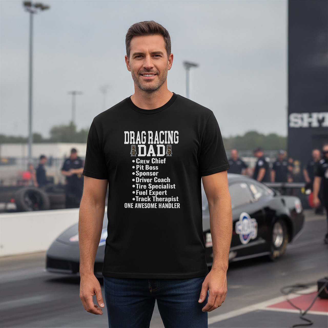 Man wearing a black 'Drag Racing Dad' t-shirt on a race track with cars and people in the background.