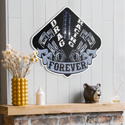 A metal sign featuring a black, white, and gray 'Drag Racing Forever' design with pistons and gears, displayed on a light gray wood-paneled wall above a wooden mantel decorated with a yellow vase of dried flowers, a small white candle, and a bear figurine.