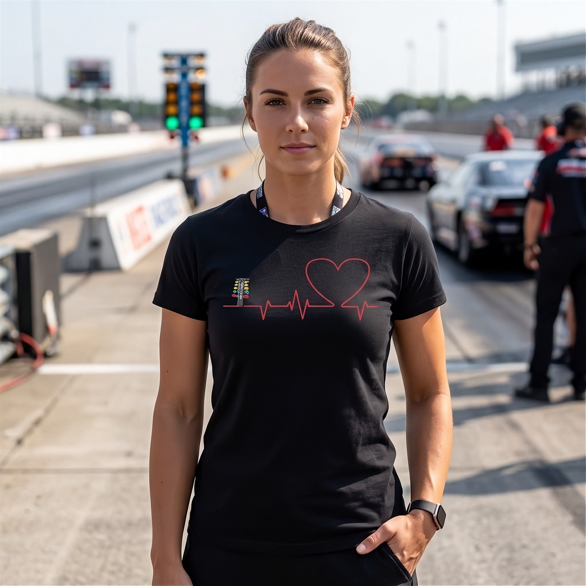 A woman stands on a racetrack wearing a black short-sleeve t-shirt with a red heart-shaped heartbeat line graphic and a small trophy icon on the chest.
