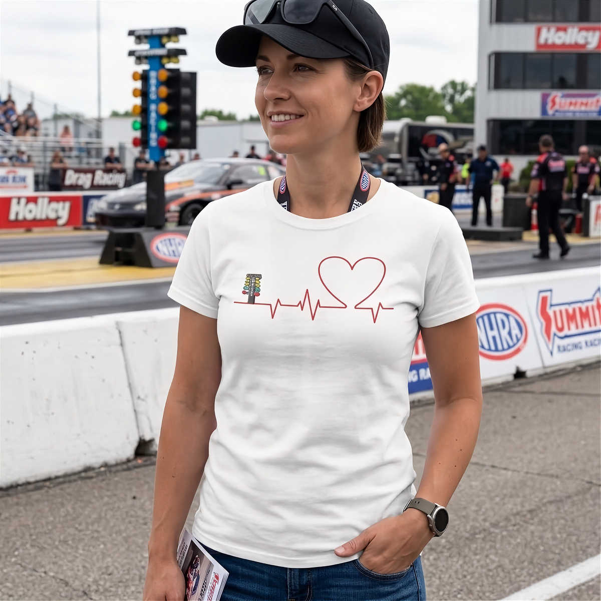 A woman wearing a white graphic t-shirt with a red heart and heartbeat line design, standing in a drag racing environment with a track and barriers in the background.