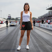 A woman stands on a racetrack wearing a white tank top and black athletic shorts with a red and white heart drag racing graphic on the left leg, paired with white sneakers. Race cars and track barriers are visible in the background.