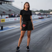 A woman stands on a racetrack wearing a black short-sleeve t-shirt and black shorts with a red and white heart graphic on the left leg. The background shows blurred racetrack grandstands, race cars, and the asphalt track.