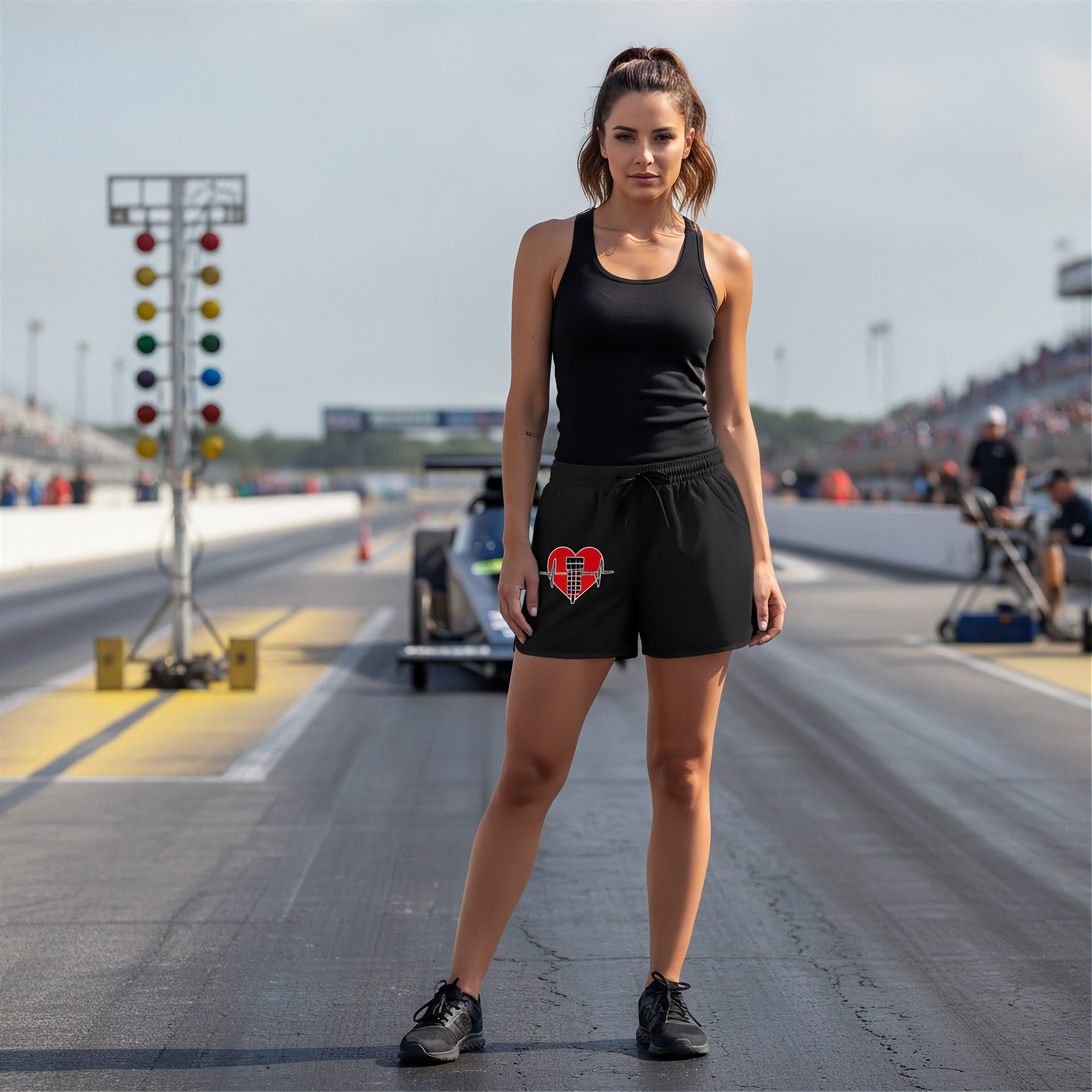 A woman stands on a racetrack wearing a black tank top and black shorts with a red heart-shaped graphic on the left leg. A drag race car is visible in the background, along with track lights and a blurred crowd in the stands.