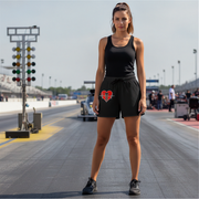 A woman stands on a racetrack wearing a black sleeveless tank top and black shorts with a red and white heart drag racing graphic on the left leg. The background shows the racetrack, starting line lights, and distant race cars under a bright sky.