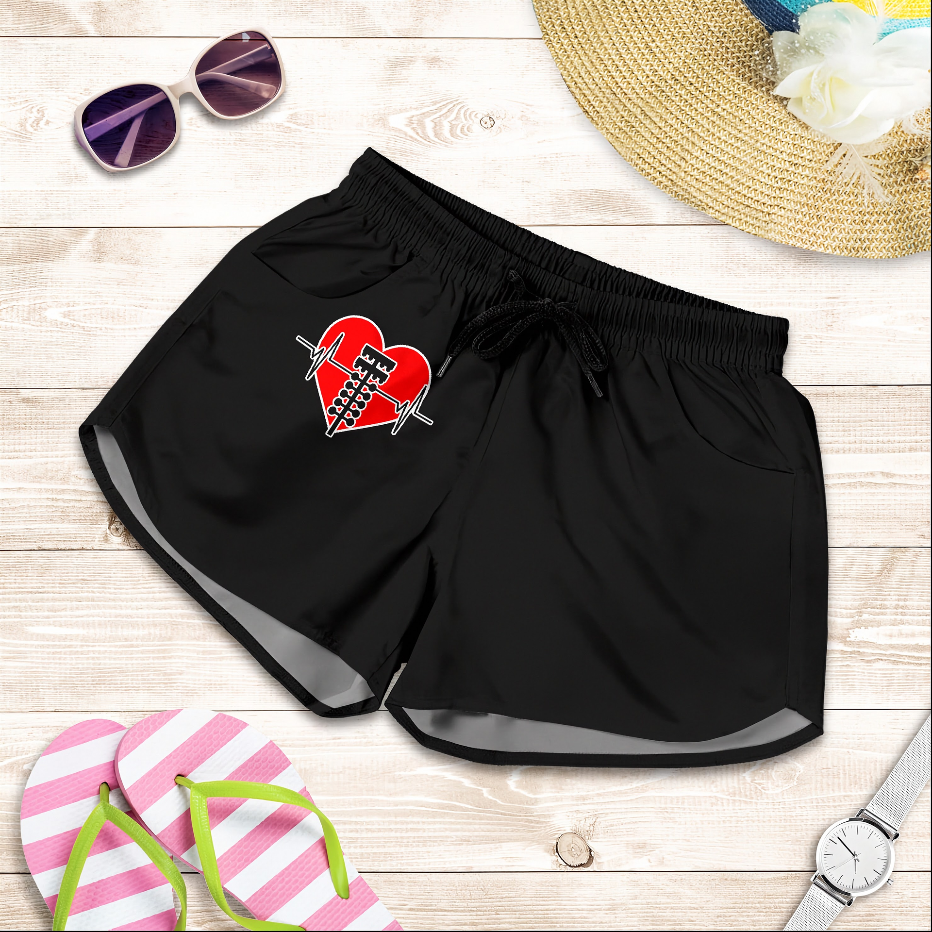 Black athletic shorts with a red heart and barbell graphic on the left leg, styled in a flat lay with sunglasses, a straw hat, pink flip-flops, and a wristwatch on a light wood background.