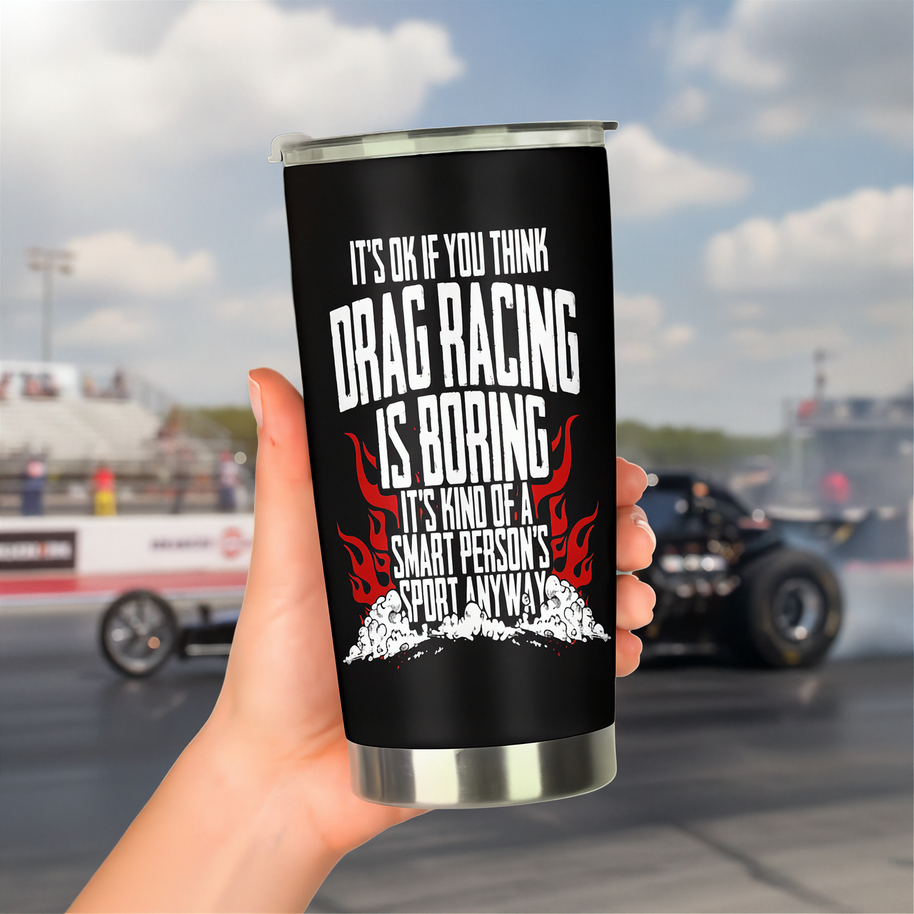 A hand holds a black insulated tumbler with a silver rim and base. The tumbler features a white and red graphic with the text 'IT'S OK IF YOU THINK DRAG RACING IS BORING IT'S KIND OF A SMART PERSON'S SPORT ANYWAY!' with flames and a drag race car in the blurred background of a racetrack.