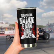 A hand holds a black insulated tumbler with a silver rim and base. The tumbler features a white and red graphic with the text 'IT'S OK IF YOU THINK DRAG RACING IS BORING IT'S KIND OF A SMART PERSON'S SPORT ANYWAY!' with flames and a drag race car in the blurred background of a racetrack.