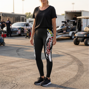 A woman stands outdoors in a paddock area wearing black athletic leggings with a large white, red, and green graphic print down one leg, paired with a black t-shirt and black sneakers.