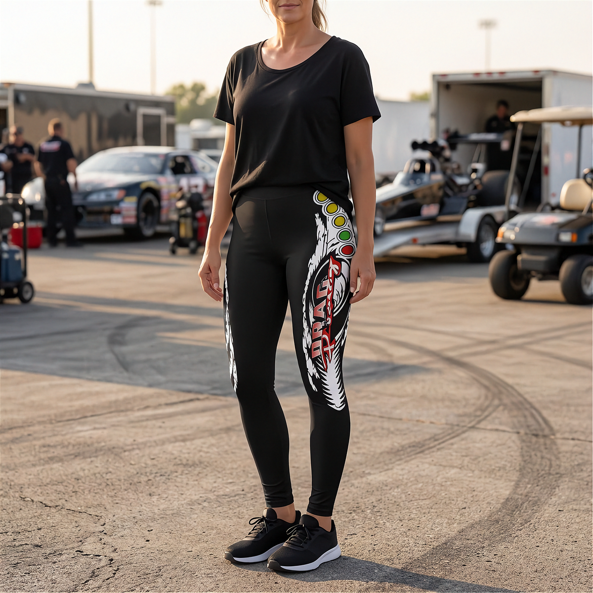 A woman stands outdoors in a paddock area wearing black athletic leggings with a large white, red, and green graphic print down one leg, paired with a black t-shirt and black sneakers.