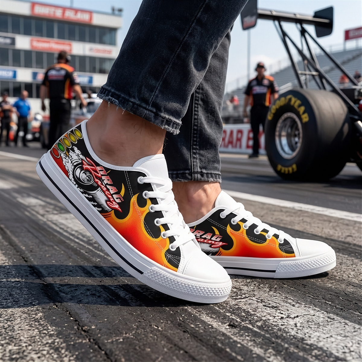 A pair of low-top canvas sneakers worn with dark jeans, featuring a vibrant flame graphic in orange, yellow, red, and black on a white background. The shoes are photographed on a racetrack with a blurred Goodyear tire and track crew in the background.