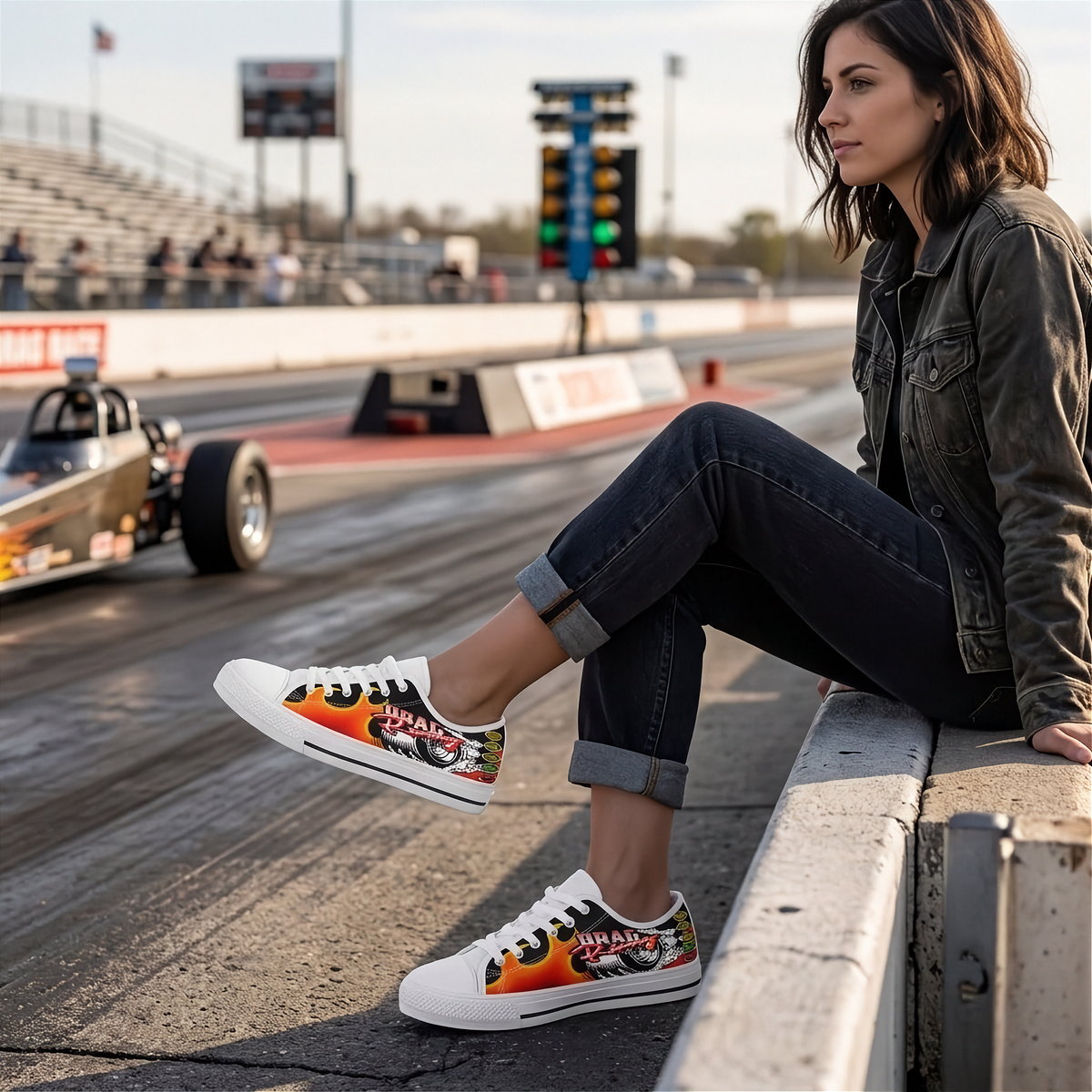 A woman sits on a concrete barrier at a drag racing track, wearing white low-top sneakers with colorful graphics. A drag race car is visible in the background on the racetrack.