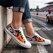 A person sitting on a concrete barrier at a racetrack wearing low-top canvas sneakers with a bold racing graphic. The shoes feature a black base with red, orange, yellow, and white flame patterns, along with a stylized racing wheel illustration and the word ' Drag Racing'.