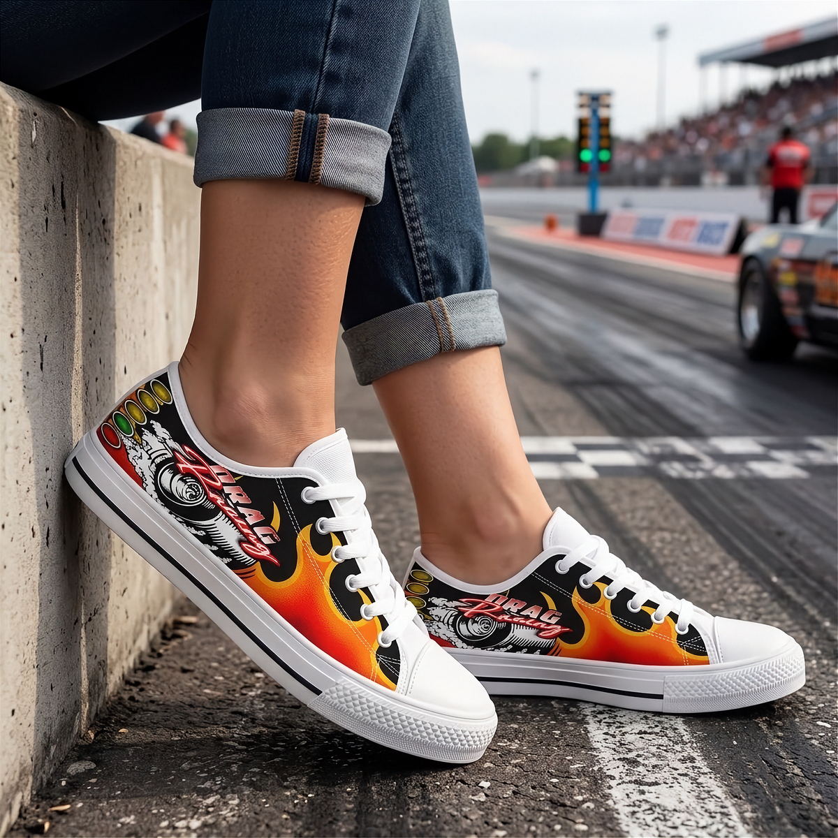 A person sitting on a concrete barrier at a racetrack wearing low-top canvas sneakers with a bold racing graphic. The shoes feature a black base with red, orange, yellow, and white flame patterns, along with a stylized racing wheel illustration and the word ' Drag Racing'.