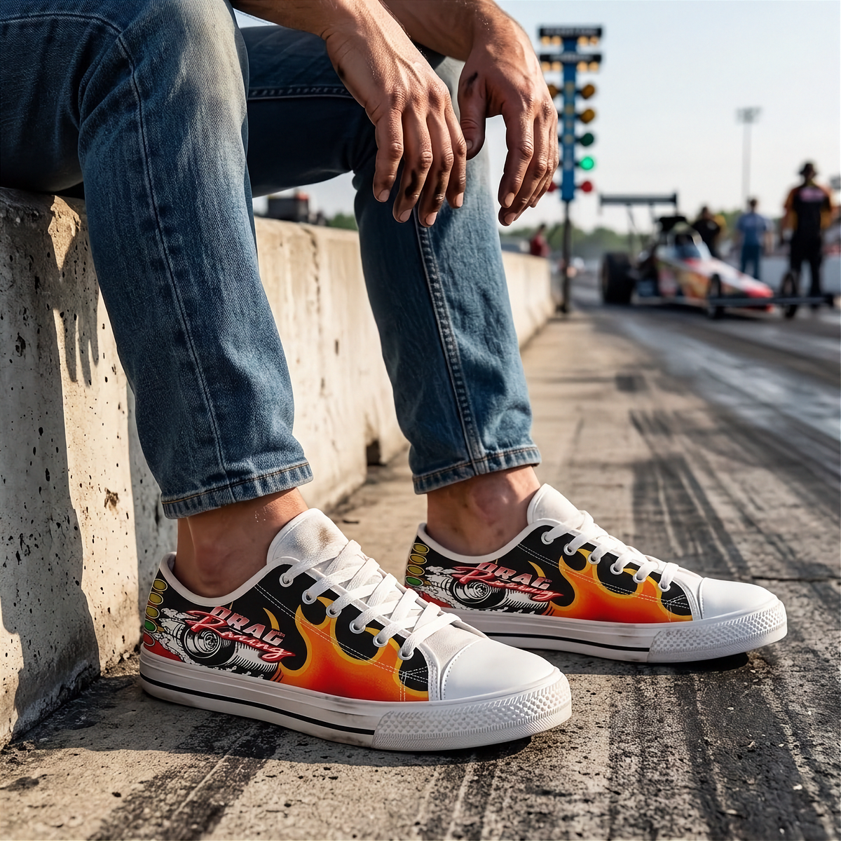 A person sitting on a concrete barrier wearing white low-top sneakers with a black, red, and orange flame graphic and a small car illustration on the side. The background shows a racetrack scene with a race car and traffic lights.